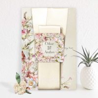 Floral metal wedding invitation propped upright next to plant in vase and rose.